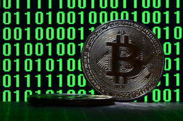 A pair of bitcoins lies on a cardboard surface on the background of a monitor depicting a binary code of bright green zeros and one units on a black background. Low key lighting