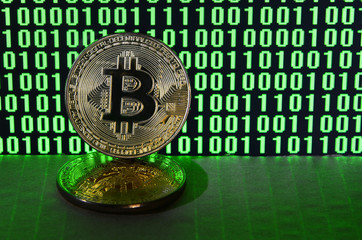 A pair of bitcoins lies on a cardboard surface on the background of a monitor depicting a binary code of bright green zeros and one units on a black background. Low key lighting