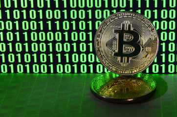 A pair of bitcoins lies on a cardboard surface on the background of a monitor depicting a binary code of bright green zeros and one units on a black background. Low key lighting