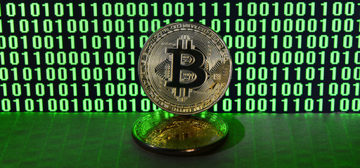 A pair of bitcoins lies on a cardboard surface on the background of a monitor depicting a binary code of bright green zeros and one units on a black background. Low key lighting