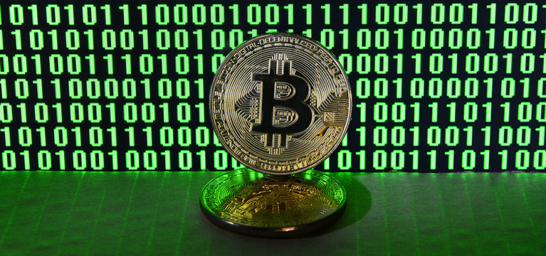 A pair of bitcoins lies on a cardboard surface on the background of a monitor depicting a binary code of bright green zeros and one units on a black background. Low key lighting