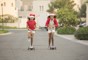 Two Cute Biracial sisters or friends plays on their scooter in the middle of the street  wearing Santa hats  - Powered by Adobe