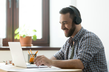 Handsome man with beard listening music in headphones and looking on screen of laptop. Young manager having a rest and playing in computer games or making video call on pc