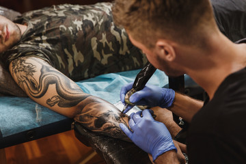 Professional tattooer artist doing picture on hand of man by machine black ink from a jar. Tattoo...