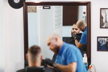 Fototapeta premium Male professional hairdresser serving client, shaving thick big beard straight razor. Ginger handsome stylish young man with short hair getting trendy haircut, black cape. Light white barber shop room