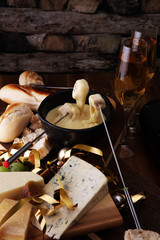 Gourmet Swiss fondue dinner on a winter evening with assorted cheeses on a board alongside a heated pot of cheese fondue with two forks dipping bread and white wine or champagne