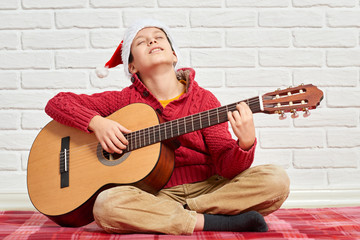 boy playing music on guitar, dressed in a red woolen sweater and santa hat, sitting on a red checkered blanket, white brick wall on background