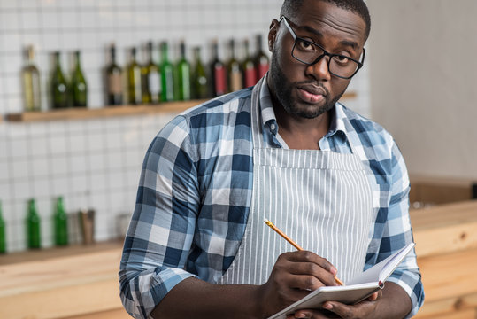 Smart Waiter. Responsible Reliable Smart Waiter Looking Serious While Standing In A Cafe And Making Notes In A Special Notebook