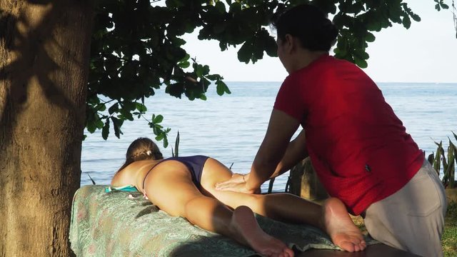 Asian woman massage therapist doing massage to girl. Summer vacation on luxury spa resort, massage procedure on the beach. Young woman enjoying a massage outside. Bali,Indonesia.