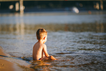 Fototapeta premium The baby boy sits on the waist in the reservoir of the river with his back and looks beyond the horizon. Concept of summer beach vacation
