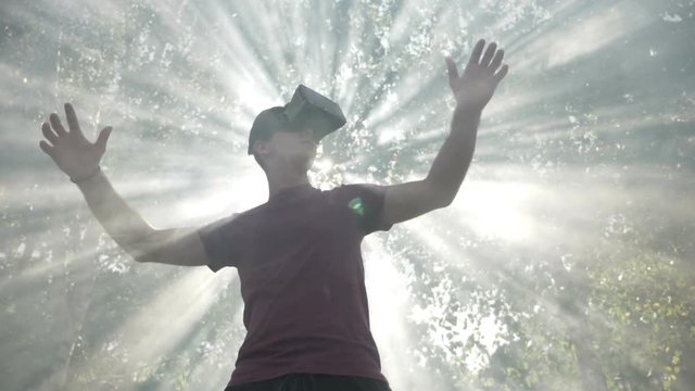 Young man with VR headset gadget immersed into virtual reality covered by light rays and waving smoke in slow motion
