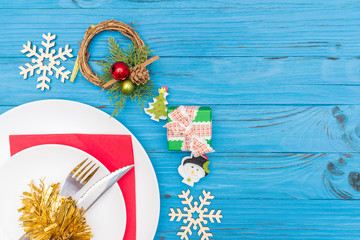 A knife and fork in white plate on red napkin decorated with a Christmas wreath wooden snowflakes and gift box on blue table