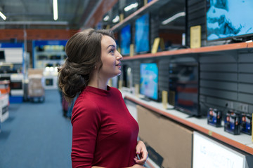 Smart modern female customer choosing large TV-sets at electronics store. She looks wondered. New screen generations.