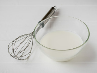 Whisk and glass bowl with milk on white wooden table.
