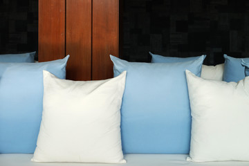 Many square shape pillows of white and light-blue colors on white sofa with the wooden pole behind