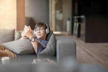 Fototapeta premium Young Asian man using smartphone for listening to music, taking a break and relaxing on sofa in living space, high school or university college student, educational concept