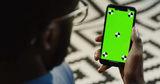 Over shoulder view on african american man holding a black smart phone vertically with green screen and tracking motion. Chroma key. Patterned background