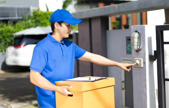 Delivery Concept - Happy Delivery Person In Blue Uniform Holding Cardboard Box Standing In Corridor Ringing Doorbell