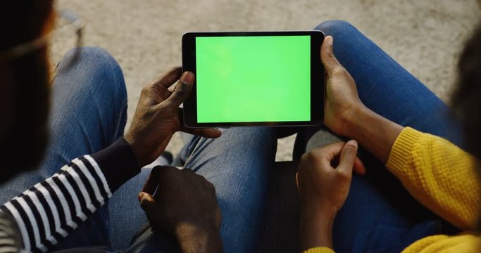 African American couple holding a black tablet device with green screen horizontally and woman scrolling and taping on it. Chroma key. Over shoulder. Close up