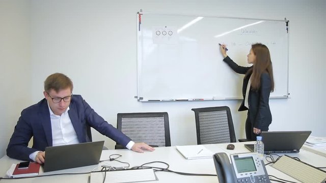 Woman writes on magnetic board, talking to man in large company. Young brunette takes notes on whiteboard with marker then chatting amiably with colleague sitting at table with laptop. Promising