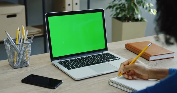 Close up of the male hands writing in the notebook with a pencil near the laptop computer with a green screen and smartphone in the office. Chroma key