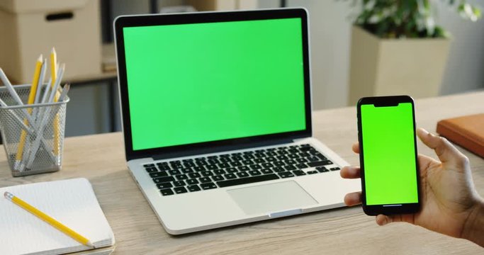 Close up of the man's hands scrolling and taping on the black smartphone with green screen and a laptop computer with green screen on the office table is on the background. Chroma key.
