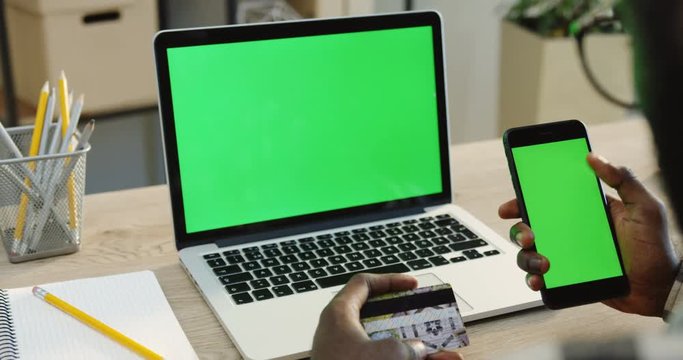 Over shoulder view on the African American man scrolling and taping on a black smartphone with green screen and holding a credit card while shopping online. Laptop computer with green screen on the