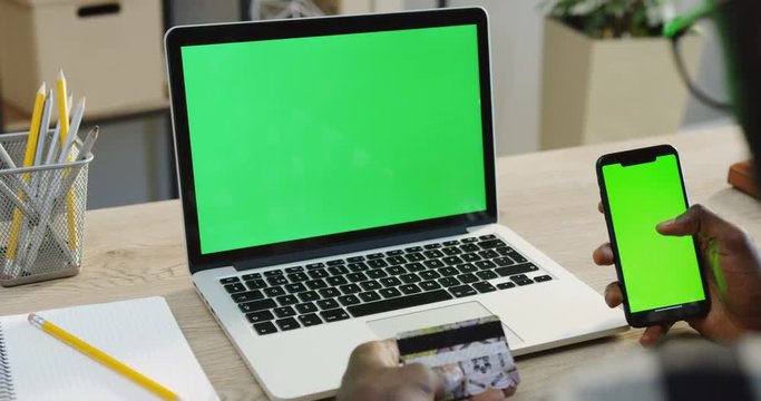 Over shoulder view on the African American man's hands scrolling and taping on a black smartphone with green screen while he holding a credit card while shopping online. Beside a laptop computer with