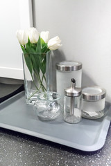 A set of modern condiment glass containers by square vase with white artificial flowers on grey tray