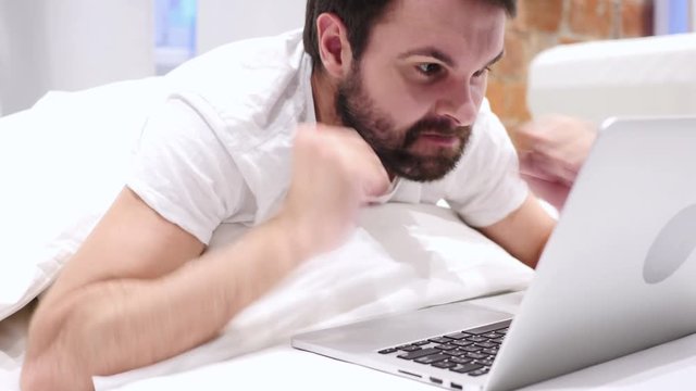 Angry Man Working on Laptop, Lying on Stomach in Bed