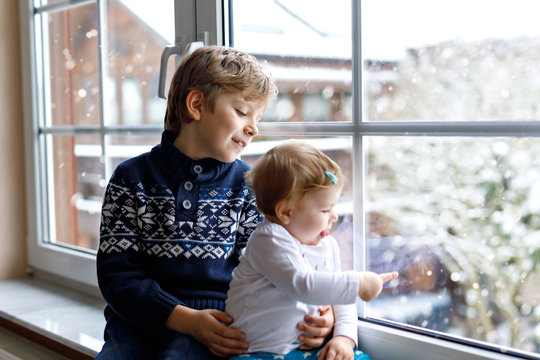 Happy Adorable Kid Boy And Cute Baby Girl Sitting Near Window And Looking Outside On Snow On Christmas Day Or Morning