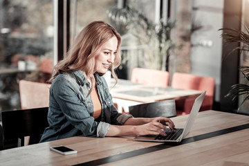 Nice place. Smart cheerful smiling student sitting in a cafe and making a report while looking at the screen of her laptop and typing