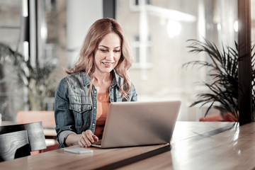 Obraz premium Convenient device. Cheerful pleasant young busy woman sitting in a comfortable cafe and looking at the screen of her modern convenient laptop while working at her report