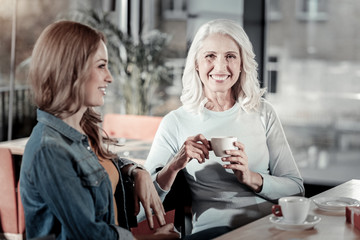 Positive women. Cheerful smiling old woman looking happy while drinking coffee and communicating with her interesting young granddaughter