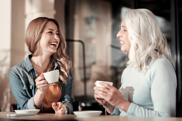Laughing. Cheerful positive senior woman and her pretty young granddaughter looking happy while sitting in a cafe and smiling at funny jokes