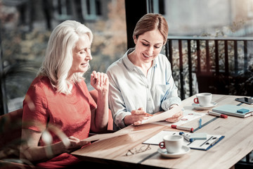 Not bad. Calm pleasant senior woman looking at the drawing of a creative young cheerful designer while sitting in a cafe with her and having a productive day