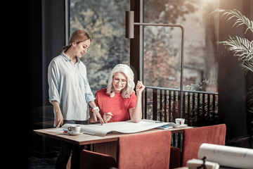 Important scheme. Cheerful friendly young designer standing near the table and pointing to the big detailed scheme while showing it to her cheerful colleague