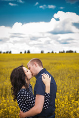 Happy young couple hugging and kissing on yellow meadow. Girl with long brunette hair in black dress in white peas, boy in blue shirt in pattern