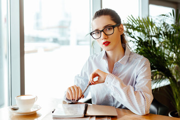 the business lady's portrait in office shirt and strict glasses. the woman sits at a table in cafe and makes entries in a notebook.