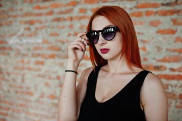 Fototapeta premium Red haired stylish girl in sunglasses wear in black, against abadoned place with brick walls.