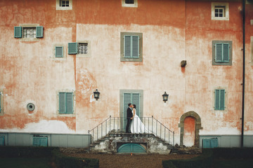 Kissing wedding couple stands on the footsteps before an orange Italian villa