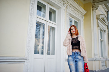 Red haired girl with red handbag posed near vintage house and speaking on mobile phone.