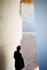 Obraz premium A young tall man in a black coat standing at the water and looks into the distance