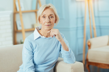 Obraz premium Pleasant pensioner. Calm pleasant aged woman feeling good while sitting in her comfortable room with her fingers touching the chin and waiting for her dear relatives to come