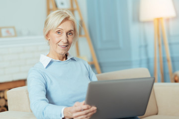 Smart woman. Enthusiastic clever senior woman looking glad while sitting on a comfortable sofa with a convenient modern laptop and smiling