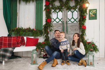 Obraz na płótnie Canvas Happy parents with little son. Child boy in sweater sitting on porch snow steps at light house with decorated in red green New Year door at home. Christmas good mood. Family and holiday 2018 concept