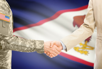 USA military man in uniform and civil man in suit shaking hands with adequate national flag on...
