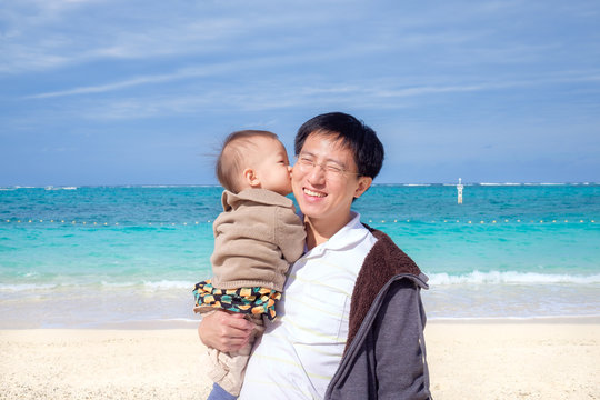 Cute little Asian 1 year old / 18 months toddler baby boy child kiss dad on beautiful white sand beach. Family travel, summer beach vacation with children, Kid & father wearing sweater, Okinawa Japan