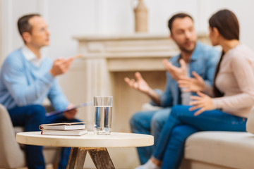 Calm water.  Selective focus of glass of crystal cold water and books placing on the wooden coffee table and couple arguing and discussing 