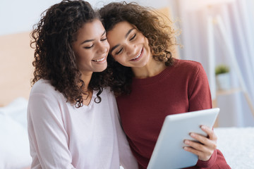 Best friends forever. Adorable sisters smiling cheerfully and hugging while sitting at home and using their touchpad together.